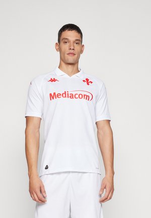 Uomo che indossa una maglia da sport bianca e pantaloncini con il logo rosso "Mediacom" e uno stemma rosso, in piedi davanti a uno sfondo chiaro uniforme.