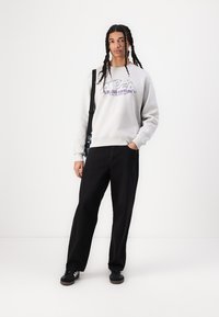 Sweatshirt gris avec le graphique "STUDIO EDITION" en violet, associé à un pantalon large noir et des baskets noires. Le mannequin a de longs cheveux en tresses.