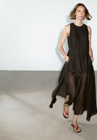 Black sleeveless dress made of lightweight fabric, featuring a loose fit, side pockets, and frayed hem, paired with brown flip-flops.