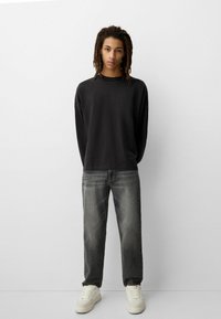 Giovane con dreadlocks che indossa una maglietta nera a maniche lunghe, jeans neri sbiaditi e sneakers bianche, in piedi contro un muro bianco semplice.