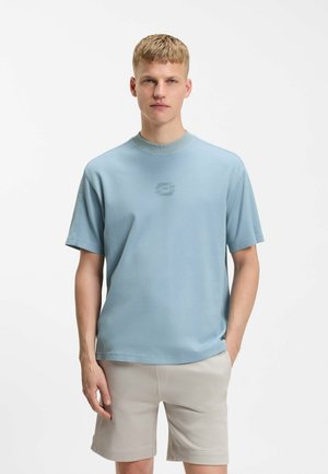 Jeune homme aux cheveux blonds courts portant un t-shirt bleu clair à manches courtes et un short beige, debout avec une main dans la poche.