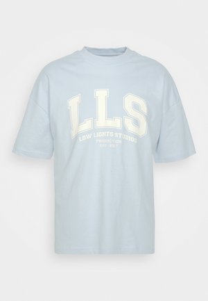 Personne portant un short LLS bleu clair et un T-shirt assorti, des chaussettes blanches et des baskets blanches, debout près d'un chariot bleu et en bois sur un sol jaune.