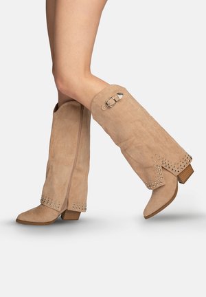 Piernas con botas de ante beige hasta la rodilla, con tachuelas a lo largo de los bordes, cremallera lateral, detalle de hebilla cerca de la parte superior y tacones bajos de madera apilada.