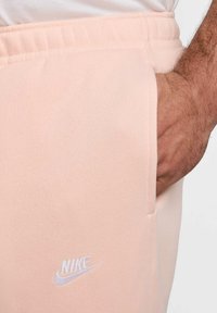 Joggers Nike de color rosa claro con una textura de tela suave, que cuentan con bolsillos laterales y un logo bordado en blanco en el lado izquierdo.