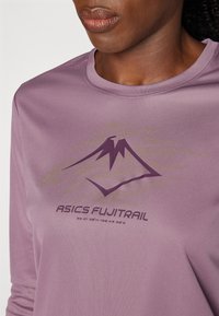 Close-up of person wearing purple Asics Fujitrail long-sleeve shirt with mountain logo and coordinates on chest.