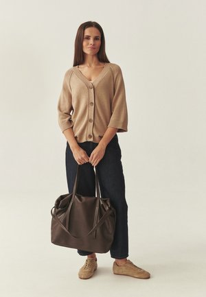 Vrouw staand in een beige vest, donkere spijkerbroek en sneakers, met beide handen een grote bruine leren shopper tas vasthoudend.