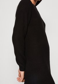 Robe noire tricotée avec une coupe ample, des manches longues et un motif texturé. Le tissu semble doux avec des accents côtelés aux poignets.