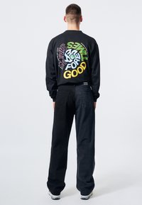 Černá dlouhá rukávová košile s barevným textem a grafikou na zadní straně. V kombinaci s volnými tmavými kalhotami a bílými teniskami. Jednoduchý design.