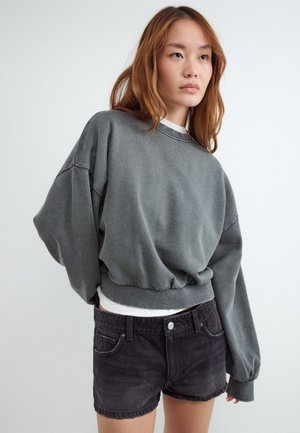 Ung kvinde med langt brunt hår iført en løs grå sweatshirt og sorte denimshorts, der står mod en ensfarvet hvid baggrund.
