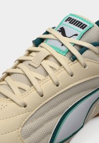 Μπεζ συνθετικό αθλητικό παπούτσι με υφασμένους πίνακες, πράσινες και λευκές λεπτομέρειες, και έντονη κορδονέτα. Διαθέτει ετικέτα με λογότυπο στη γλώσσα του παπουτσιού.