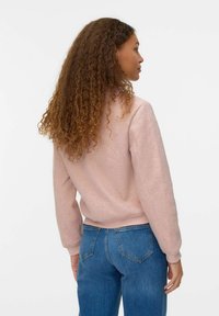 Mujer con cabello largo y rizado, vestida con un suéter rosa claro y jeans azules, de pie y mirando ligeramente hacia la derecha contra un fondo blanco.