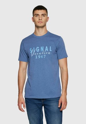 Ung mand med kort mørkt hår iført en blå T-shirt med teksten "Signal Vacation 1967" og blå jeans, står foran en ensfarvet baggrund.