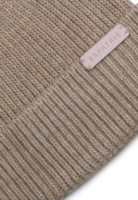 Beige neulepipo, jossa on ribbikuvio. Sivussa on nahkapehmeä etiketti, jossa lukee "EXPATRIÉ". Pehmeä materiaali varmistaa napakan istuvuuden.