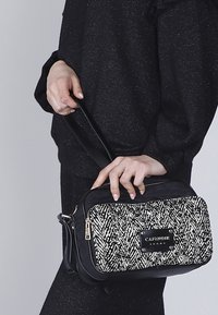 Bolso bandolera negro con un patrón de espiga blanco y negro texturizado, que presenta una cremallera dorada y un acento de logo sobre un material liso.