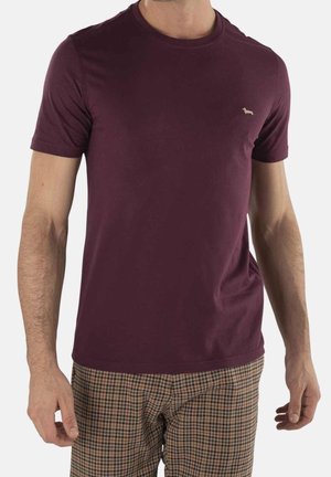 Mężczyzna w bordowej koszulce z krótkim rękawem z małym logo psa na piersi i brązowych, kratkowanych spodniach.