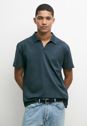 Giovane uomo in piedi con le mani dietro la schiena, indossa una camicia scura a maniche corte con colletto e jeans azzurri chiari su sfondo bianco.
