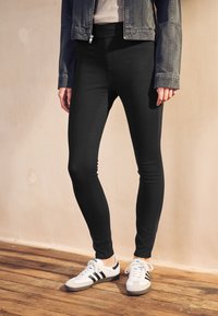 Juodos tamprios, liekną siluetą pabrėžiančios tamprės, derinamos su baltai juodais sportiniais bateliais, medinė grindų danga, neutralaus fono.