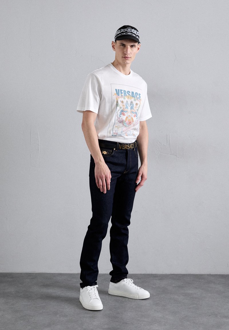 Witte t-shirt met een kleurrijke Versace grafische print, donkere denim jeans en witte sneakers, aangevuld met een zwart-wit patroontje pet.