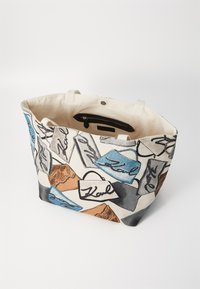 Canvas tote bag featuring a geometric multi-color design with blue, brown, and black accents, and printed logos. Zippered inner pocket included.