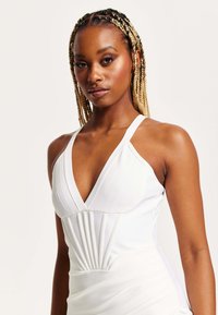 White sleeveless dress with a deep V-neck, pleated detailing at the front, and a fitted silhouette. Smooth, stretchy fabric.