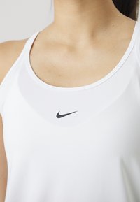 Top atlético blanco sin mangas con escote redondeado, que presenta un pequeño logo negro de Nike en la zona del pecho. Textura suave, diseño minimalista.