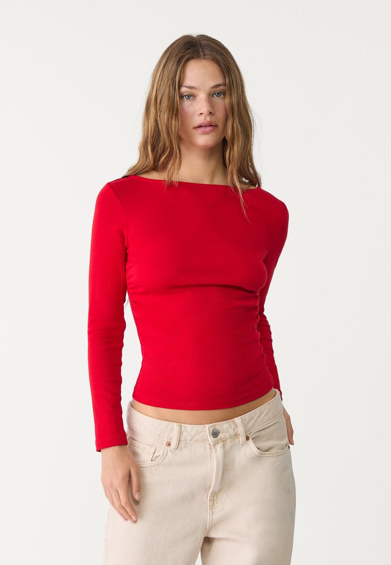 Rode langekleurige top met een bootkraag, aansluitend ontwerp en een gladde textuur. Gekozen in combinatie met lichtbeige high-waisted jeans.