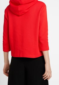 Sudadera corta roja con un patrón texturizado, mangas tres cuartos y capucha con cordón, combinada con pantalones de pierna ancha negros.