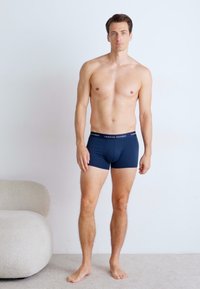 Sötétkék pamut boxer alsónadrág, testhezálló fazonnal, kontrasztos derékpánttal, amelyen a "TOMMY HILFIGER" felirat olvasható. A modell egy bézs szőnyegen áll.