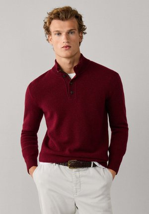 Jeune homme aux cheveux blonds bouclés portant un pull bordeaux boutonné, une chemise blanche, un pantalon beige et une ceinture marron tressée, les mains dans les poches.