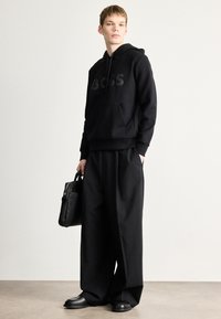 Μαύρο φούτερ με κουκούλα με το λογότυπο "BOSS", σε συνδυασμό με μαύρο παντελόνι φαρδιάς γραμμής. Το μοντέλο κρατά μια μικρή μαύρη τσάντα και φορά μαύρα παπούτσια. Απλό, μονόχρωμο στυλ.