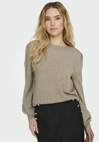 Beige stickad tröja med ribbad textur och breda ärmar, kombinerad med en svart kjol med dekorativa knappar på sidan.