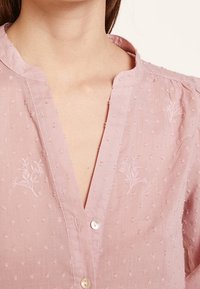 Blusa rosa claro con un tejido texturizado que presenta puntos elevados, un escote en V y sutil bordado floral. Tiene un cierre de botón.