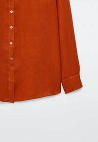 Chemise en lin couleur rouille avec manches longues, devant à boutons, col et un seul poignet. Présente un tissu doux et texturé.