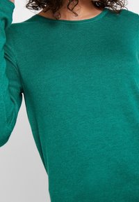 Pull en tricot côtelé couleur sarcelle avec un col rond et des manches longues, mettant en valeur une texture douce et un design ajusté.