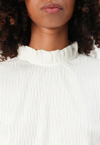 Donna vista di spalle con il viso e il collo vestiti con una blusa bianca a pieghe con colletto alto, capelli ricci castano scuro che incorniciano il viso.