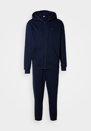 Sweat à capuche zippé bleu marine avec poches sur le devant et pantalon de jogging assorti à coupe fuselée, présenté sur un fond clair uni.