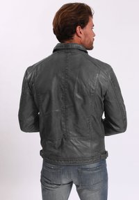 Manteau en cuir gris au toucher lisse, caractérisé par un col montant, une coupe ajustée et de subtils détails de couture. Porté sur un jean bleu.