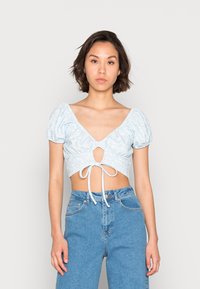 Top cropped azul claro com mangas bufantes, decote em V e um detalhe frontal em forma de buraco amarrado com um cordão. Padrão floral suave.