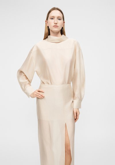 Femeie cu păr blond drept poartă un top bej cu mâneci lungi și o fustă asortată cu o deschizătură frontală, stând în picioare pe un fundal clar simplu.