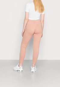 Kvinna med långt blont hår som bär en vit kort t-shirt, ljusrosa joggers och vita Nike-sneakers stående mot en neutral vägg.