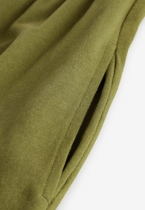 Tissu vert olive avec une texture côtelée, comportant un détail de poche latérale. Le matériau semble doux et se drape délicatement.