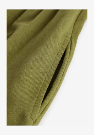 Tela de color verde oliva con una textura acanalada, que presenta un detalle de bolsillo lateral. El material parece suave y se cae con delicadeza.