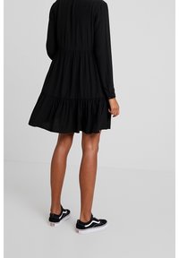 Robe noire à manches longues avec un ourlet à volants en étages, confectionnée en tissu doux. Associée à des baskets noires avec des accents blancs.