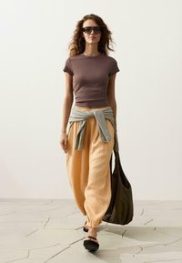 Femme aux cheveux ondulés portant des lunettes de soleil, un t-shirt marron ajusté, un pantalon beige ample, un pull rayé noué autour de la taille, et portant un sac fourre-tout sombre.