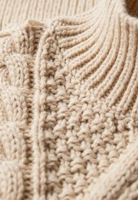 Beżowy sweter z dzianiny z ribbowymi i kablowymi teksturami, z wysokim kołnierzem i skomplikowanymi wzorami na całej powierzchni materiału.