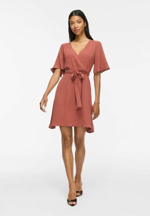 Femme en robe portefeuille couleur rouille à manches courtes avec ceinture à la taille, portant des talons ouverts noirs, debout devant un fond blanc.