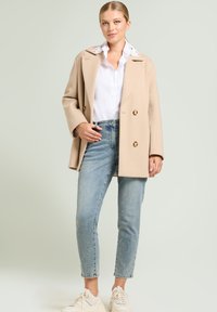 Abrigo beis de doble botonadura con grandes botones dorados, utilizado sobre una camisa blanca, combinado con jeans de un tono azul claro y zapatillas blancas.