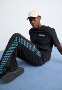 Svart t-skjorte og bukser med turkise striper. Modellen har på seg en beige caps, sitter på en lys blå overflate, med svarte sneakers.