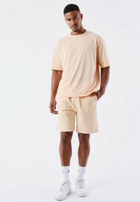 T-shirt corta a maniche corte color pesca con logo goffrato, abbinata a pantaloni corti pesca coordinati. Il modello indossa sneakers bianche e calzini a coste.