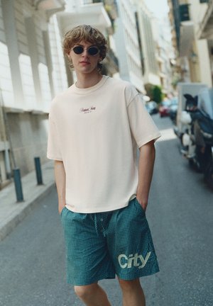 Ung person iført runde solbriller, en hvid T-shirt og blåternede shorts med teksten "City", stående på en bygade med køretøjer.